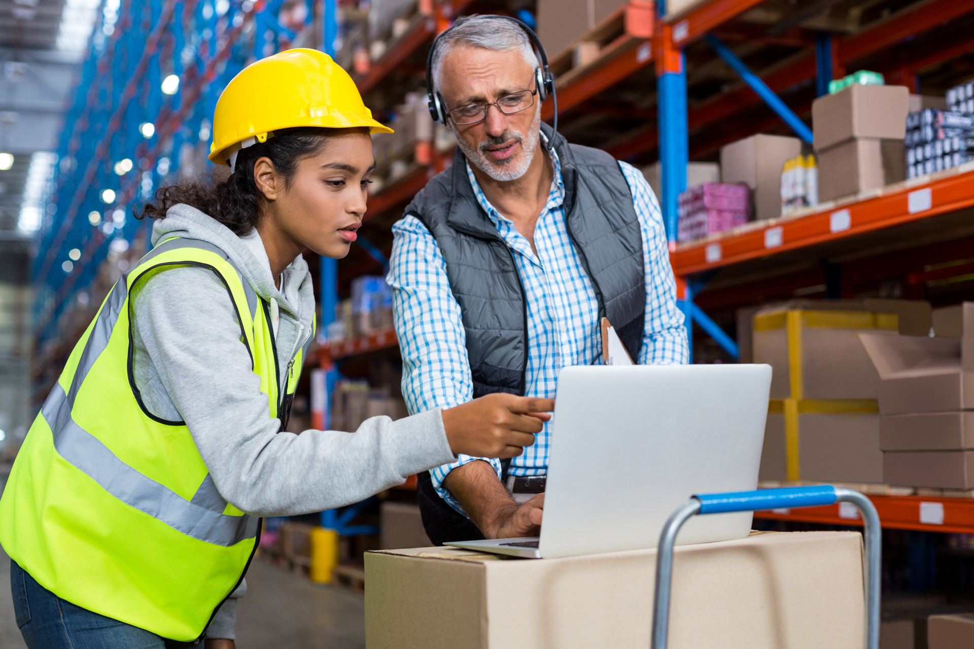 Warehouse manager and female worker in a safety helmet and vest discussing information on a laptop, surrounded by boxes and shelves in a warehouse.